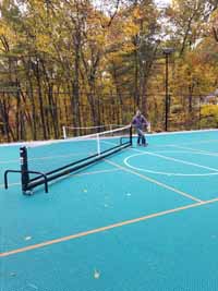Demonstrating deployment of portable yet durable net system to center of large basketball multicourt in Bolton, MA. Court was ordered with lined for pickleball. Portable nets roll easily then lock down, giving option of net sports like tennis on demand without permanent posts or nets.