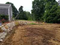 Charcoal and titanium Cape Cod backyard basketball court in Barnstable village of Marstons Mills, MA. Before picture with the ground being prepared to allow a base to be installed. Compare to after picture to right.