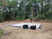 Green and red residential basketball court in Middleborough, MA. Shown here: Completed concrete base and installed goal system, with pallets of court tiles awaiting installation.