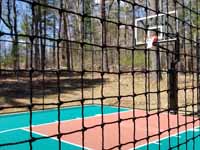 Residential basketball court in Norwell, MA, including goal system, mesh fencing, and an emerald green and rust red tile sport surface. Some trees and stumps were removed to make room in the yard and provide easy work access. This is a view of part of the court through the mesh fence.