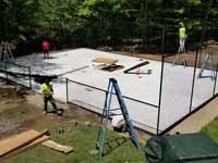 Residential backyard basketball court on fresh concrete base in stalled in Pepperell, MA, featuring fencing and an emerald green, titanium, and rust red sport tile surface. Shown here with the concrete base cured and the fence being installed.