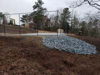 Multicourt for pickleball and basketball, fenced on three sides, on a hillside backyard in Plymouth, MA. View from the end opposite the gate, showing concrete base ready to have the tiles installed from the pallet in background.
