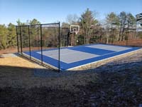 Hillside court primarily for pickleball, accessorized with a basketball goal and fencing, in Plymouth, MA. A look at the completed blue and grey court, in which adjacent stones for drainage are also visible.