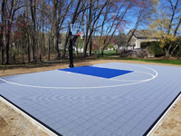 Angled view from front left corner of ice and royal blue backyard basketball court in South Hadley, MA. Small home court in black and red tile surface with custom logo designed from letters E and P.