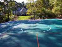 View of much of large emerald green and titanium backyard basketball court in Bolton, MA, with orange pickleball lines, for use with portable net, in foreground.