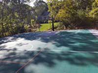 View of one end of large emerald green and titanium backyard basketball court in Bolton, MA, mottled with sun and tree shadows.