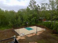 Overview of sand and green backyard basketball court in Londonderry, NH, featuring custom logos and text, optional net for additional court sports like tennis or volleyball, optional lighting for night play, and rebound fence.