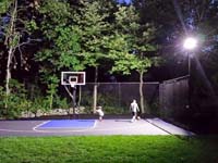 The same back yard corner shown to the right, with a completed residential basketball court shown in use under lights at night in West Bridgewater, MA.