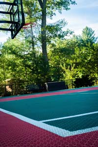 Fairly close look at the red and green tile colors at one end of a backyard basketball court with multicourt net for tennis or volleyball, adjacent in-ground trampoline, and lighting system in Pembroke, MA.