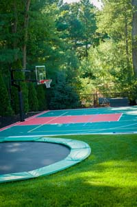 In-ground trampoline in foreground of partial view of full red and green backyard basketball court with net for tennis or volleyball n Pembroke, MA.