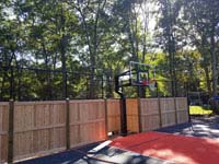 Graphite and orange residential basketball court in Walpole, MA, highlighting goal and custom fencing combining cedar and more traditional court containment fence. Also visible, a section of rebound fence without the wooden portion.
