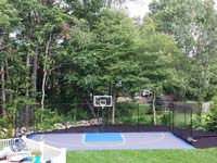 Finished view of graphite and light blue basketball court in West Bridgewater, MA, picturesque in the finished landscape and tree-laded suburban backdrop.