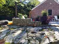 This shows the stone wall and patio area, plus part of the yard where the court will go, prior to Chestnut Hill, MA transformation of a tight backyard space into a slate green basketball court with custom containment net fencing atop wooden fence.