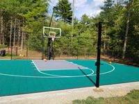View from just left of front center of court, toward hoop and right rear of court, showing lighting, hoop system, and post for optional, adjustable net for tennis or pickleball, in Dover, MA.
