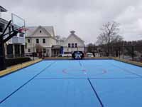 Looking down the length of a combined hockey and basketball court in Hingham, MA. Shows fence, hockey net and basketball goal options, court tiles in shades of blue, and pleasing choice of line colors.