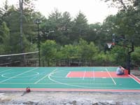 Professional installation in progress on a large backyard basketball court in Pembroke, MA, including accessories like a mid-court tennis and volleyball net with adjustable height, and lights for night games.