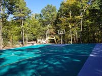 Angled view of about two-thirds of large emerald green and titanium backyard basketball court in Bolton, MA, with construction all but complete.