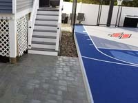Small blue and grey basketball court with custom red H logo by existing pool in Braintree, MA, looking toward hoop end and featuring associated new patio by deck and bulkhead.