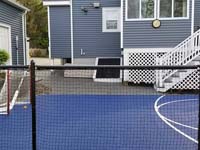 View through back fence of blue and grey basketball court in Braintree, MA, along with associated hardscapes.
